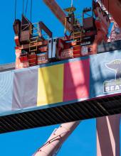 A shipping container with a Belgium flag banner hangs from a crane at Terminal 46 in Seattle.